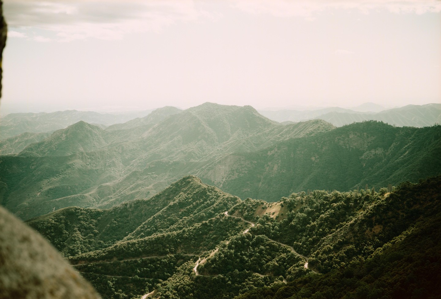 A winding road cuts through a lush, green mountain landscape under a hazy sky.