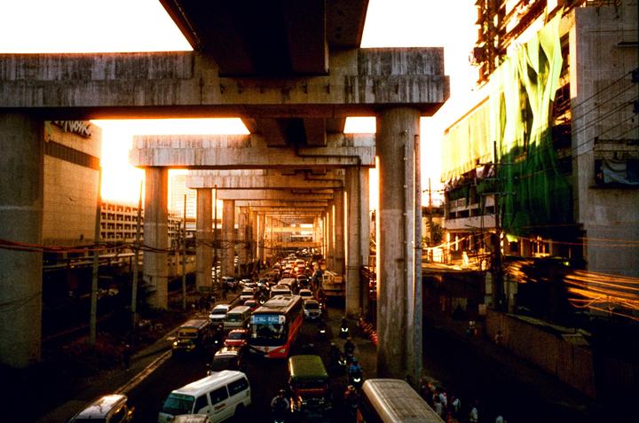 A busy street with heavy traffic is visible under a series of concrete overpasses during golden hour.