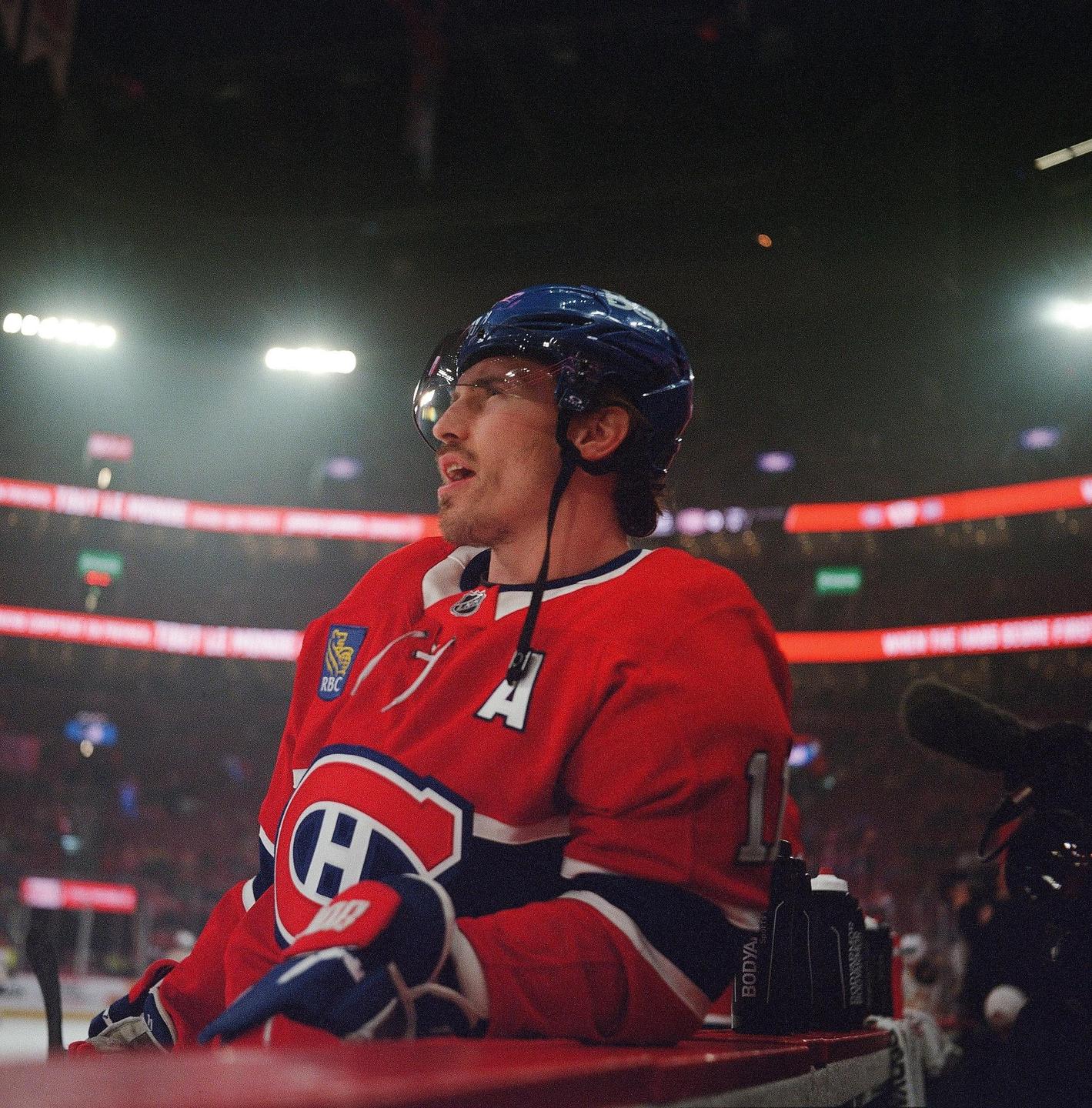 A hockey player in a red jersey and blue helmet sits on a bench during a game.
