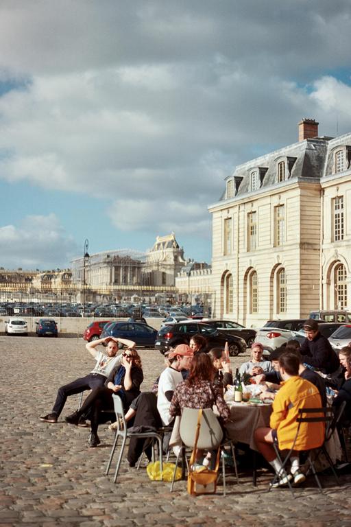 A group of people are seated around a table on a cobblestone street with a large building in the background.