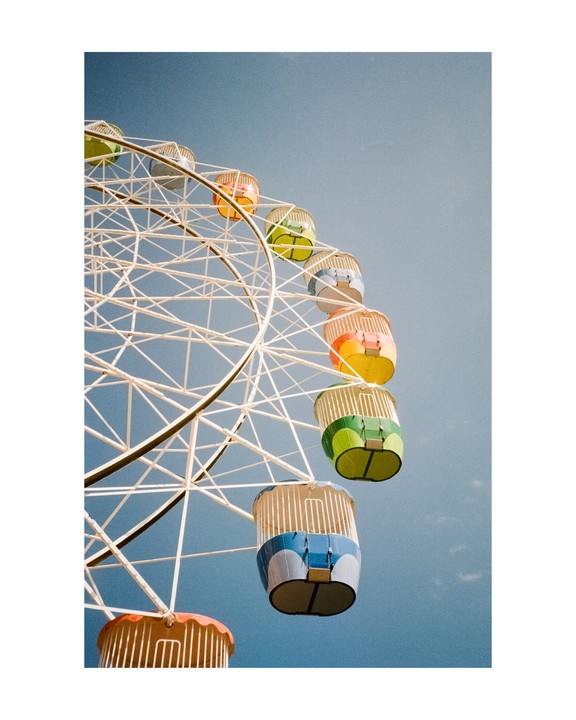 A low-angle view shows a white Ferris wheel with colorful gondolas against a clear blue sky.