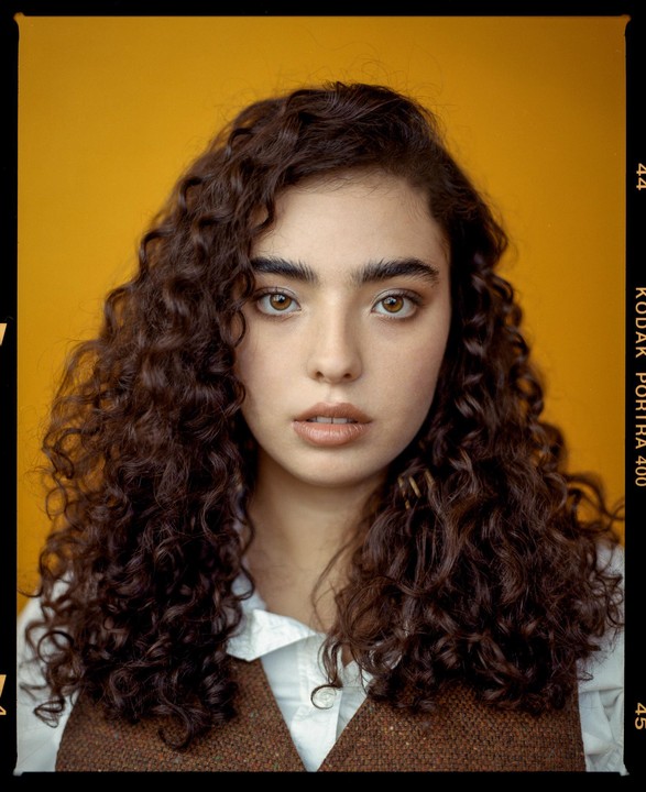 A woman with curly hair looks directly at the viewer against a yellow background.