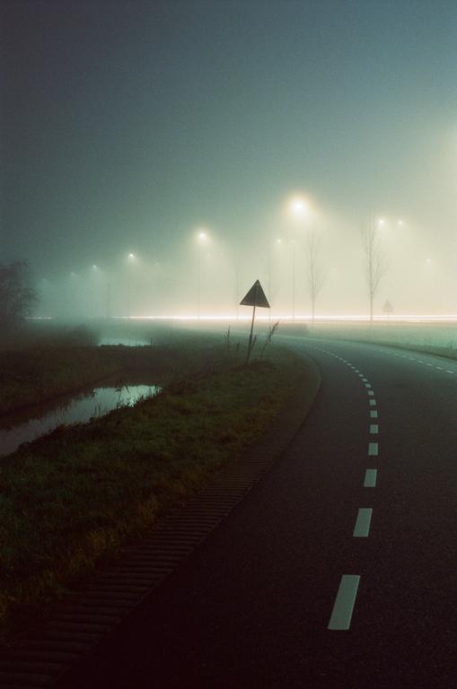 A road sign stands beside a winding road with streetlights illuminating a misty evening.