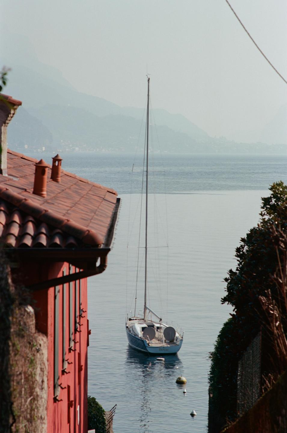 A sailboat floats on a lake with mountains in the background, framed by a red building and foliage.