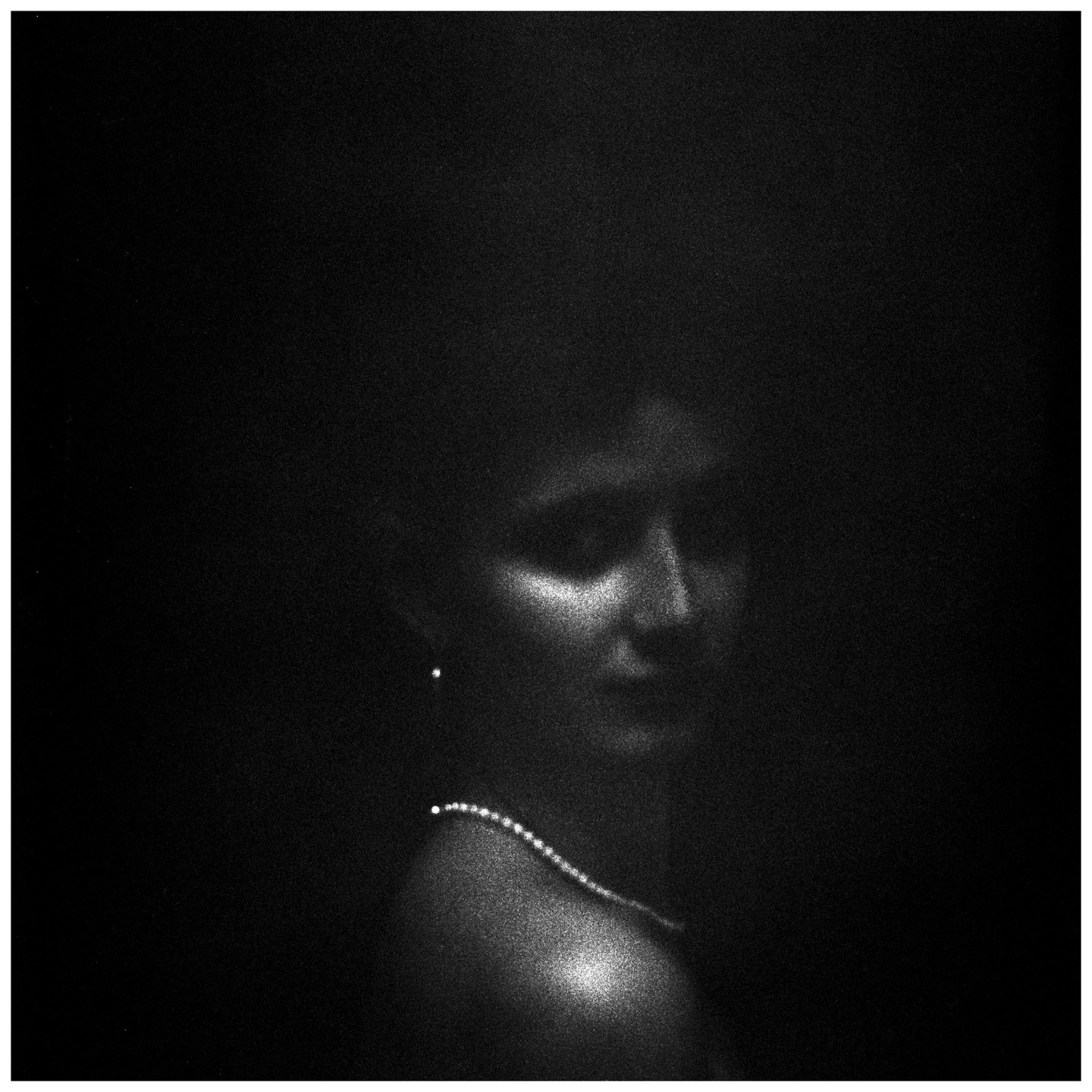 A woman's face and shoulder are dimly lit in a dark, grainy, black and white portrait, with a pearl necklace and earring visible.
