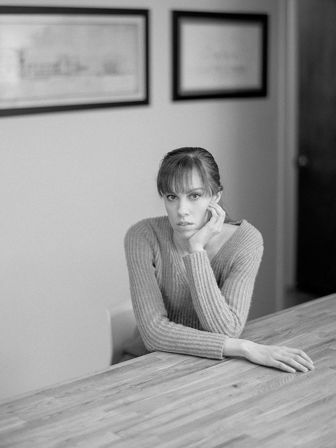 A woman with bangs and a ribbed sweater sits at a wooden table with her chin resting on her hand.