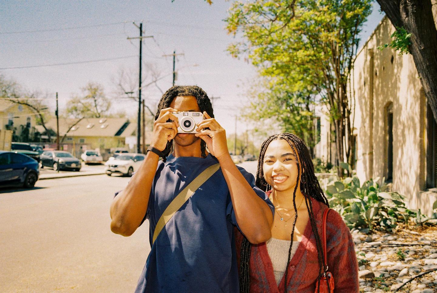 A man holds a camera to his face while a woman smiles next to him on a sunny street.