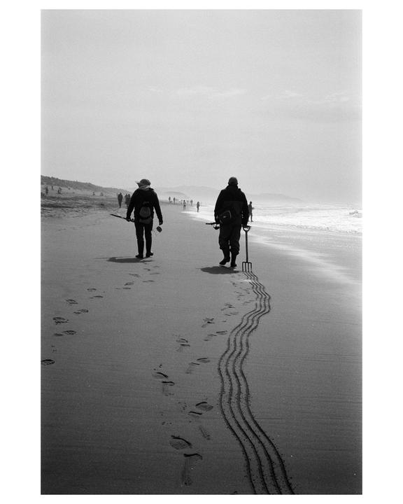 Two people walk along a beach, one dragging a rake that leaves wavy lines in the sand.