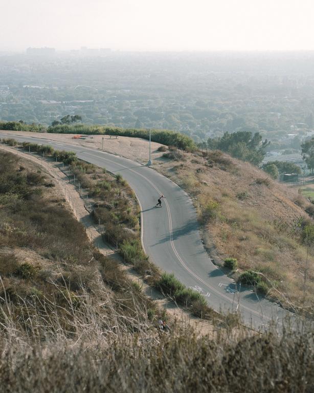 A person on a skateboard descends a winding road on a hill overlooking a hazy city.