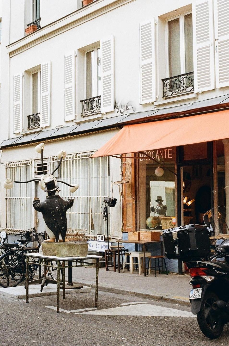 A street in front of a building with an orange awning features a statue of a man in a top hat and a scooter.