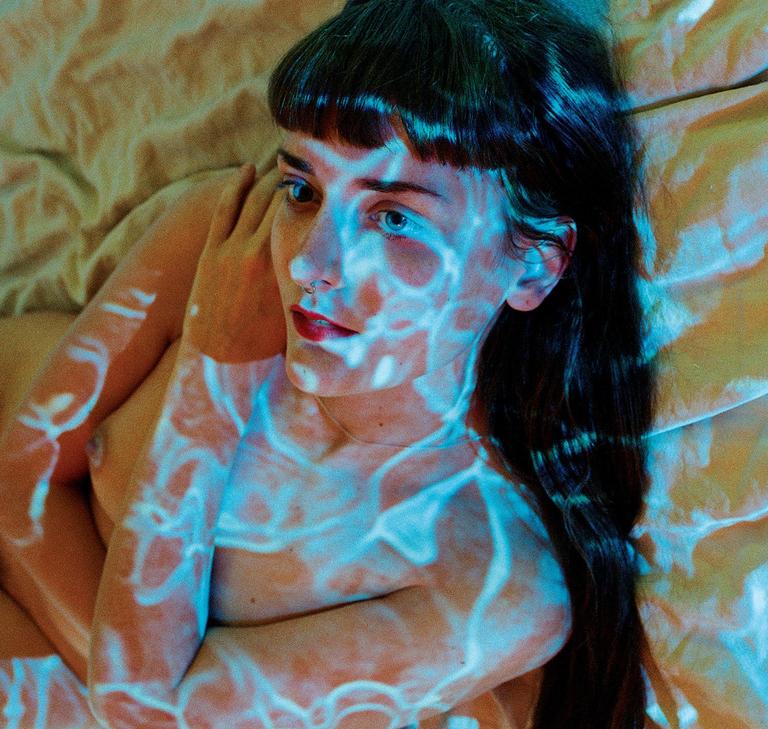 A woman with long dark hair and bangs lies on a bed with blue light patterns projected onto her body.