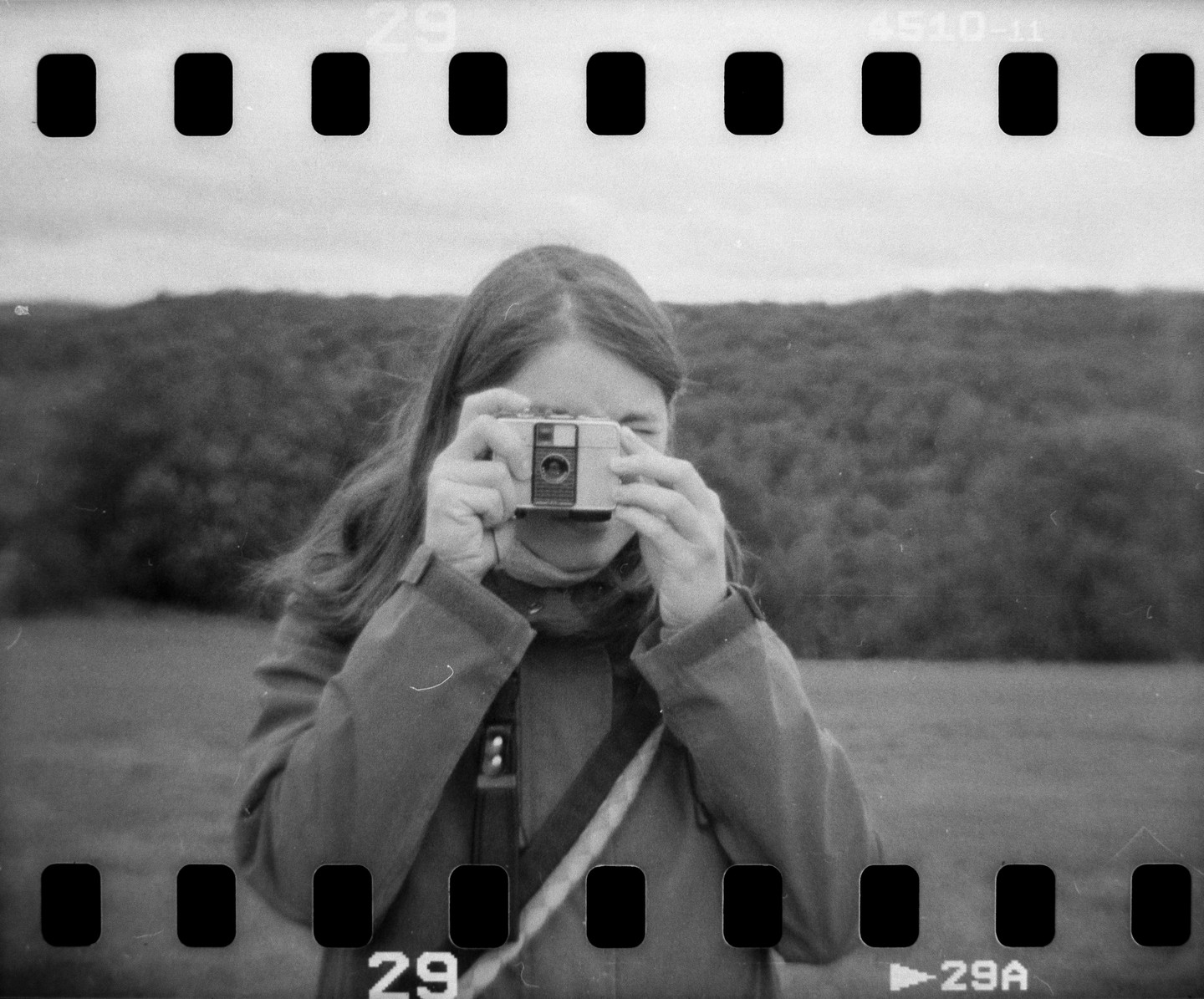 A woman holds a camera to her face, with a forest in the background.