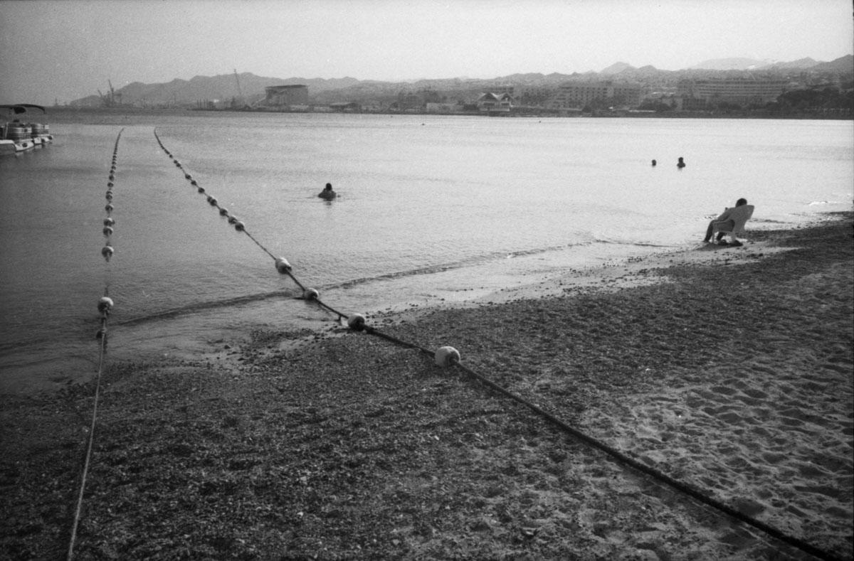 A person sits on a chair at the edge of a beach with two people swimming in the water and a city in the distance.
