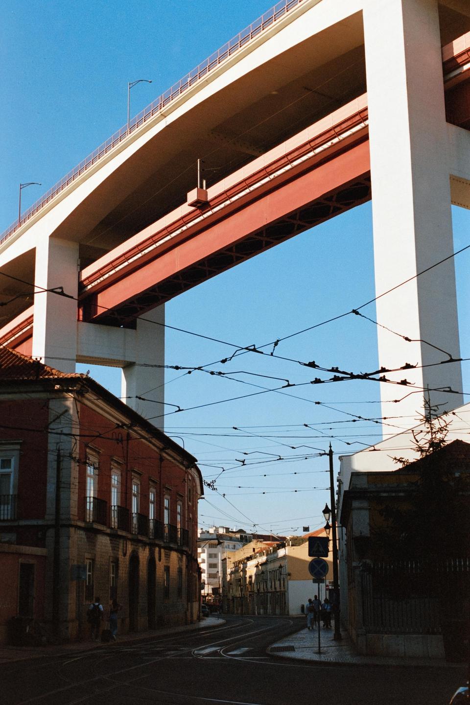 A large bridge with red and white supports spans over a street with buildings and overhead wires.