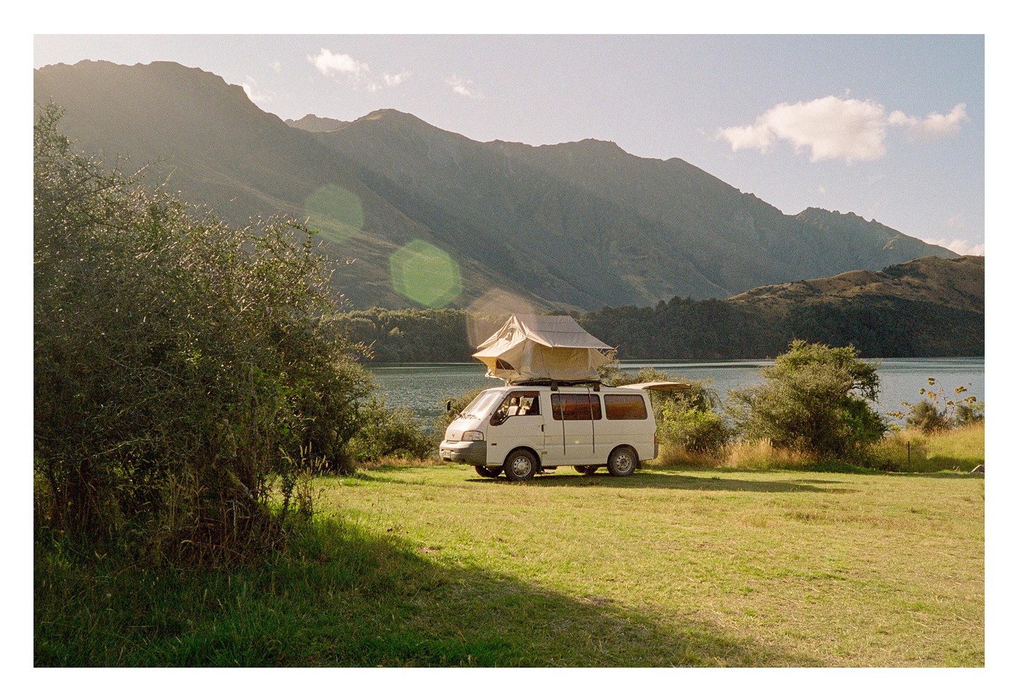 A white camper van with a tent on its roof is parked on a grassy field next to a lake, with mountains in the background.
