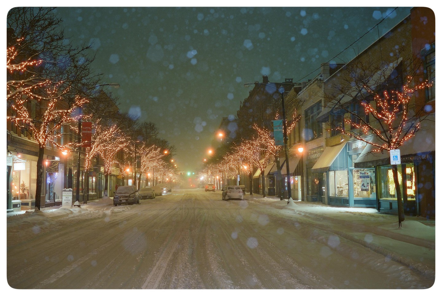 A snow-covered street at night is lined with trees adorned with string lights and buildings with illuminated storefronts.