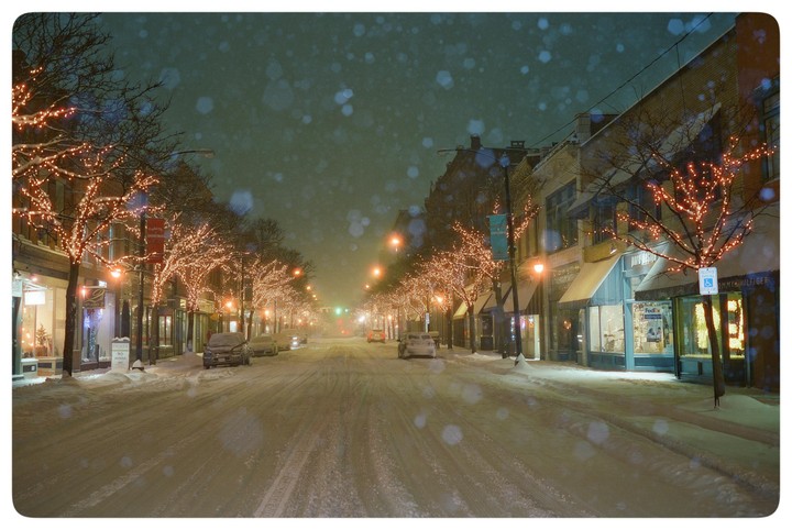 A snow-covered street at night is lined with trees adorned with string lights and buildings with illuminated storefronts.