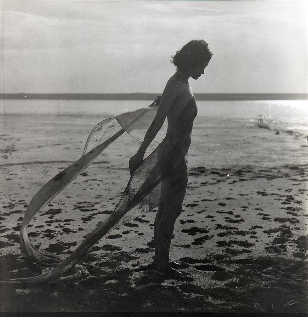 A woman stands on a sandy beach with a flowing piece of fabric around her body, looking down at the ground.