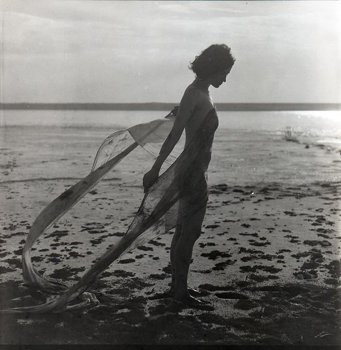 A woman stands on a sandy beach with a flowing piece of fabric around her body, looking down at the ground.