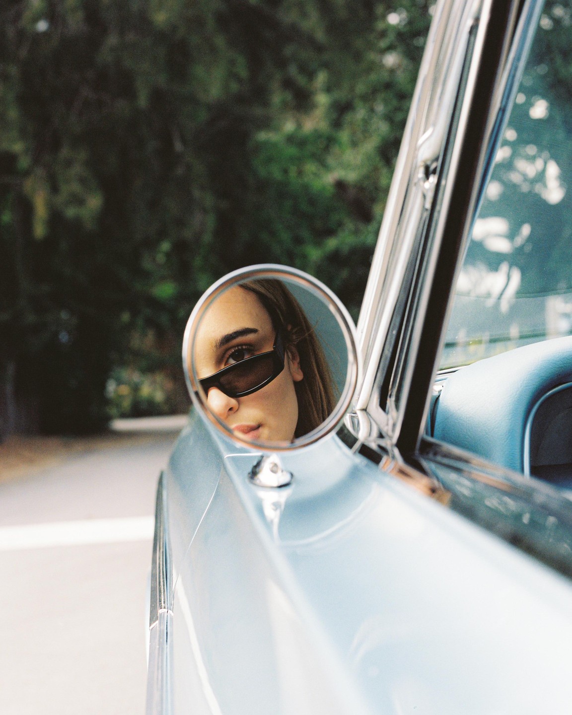 A woman wearing sunglasses is reflected in the side mirror of a light blue car.