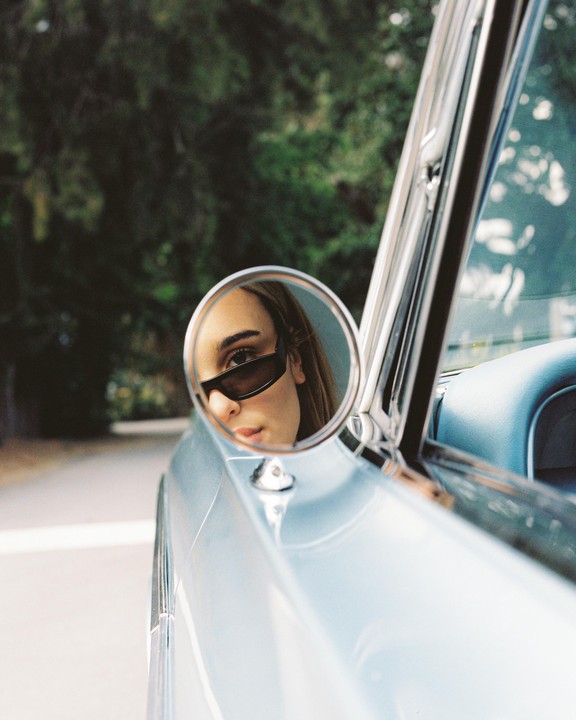 A woman wearing sunglasses is reflected in the side mirror of a light blue car.