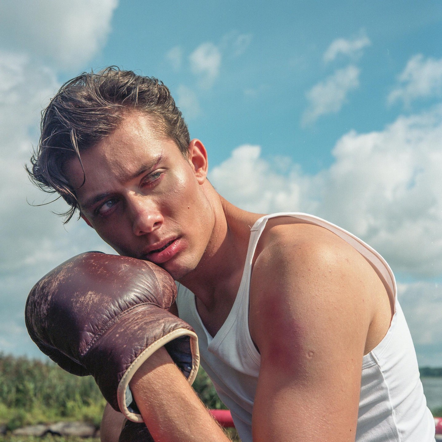A man wearing a white tank top and a boxing glove rests his head on his arm while looking away from the camera.
