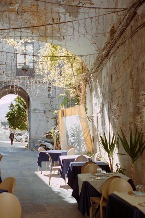 A restaurant patio with tables and chairs is set up under an archway adorned with string lights, with a person walking in the distance.
