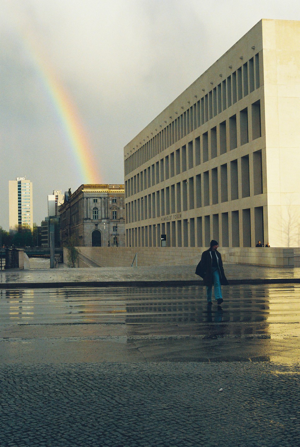 A man walks across a wet street with a rainbow in the sky over buildings.