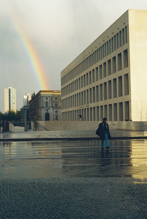 A man walks across a wet street with a rainbow in the sky over buildings.
