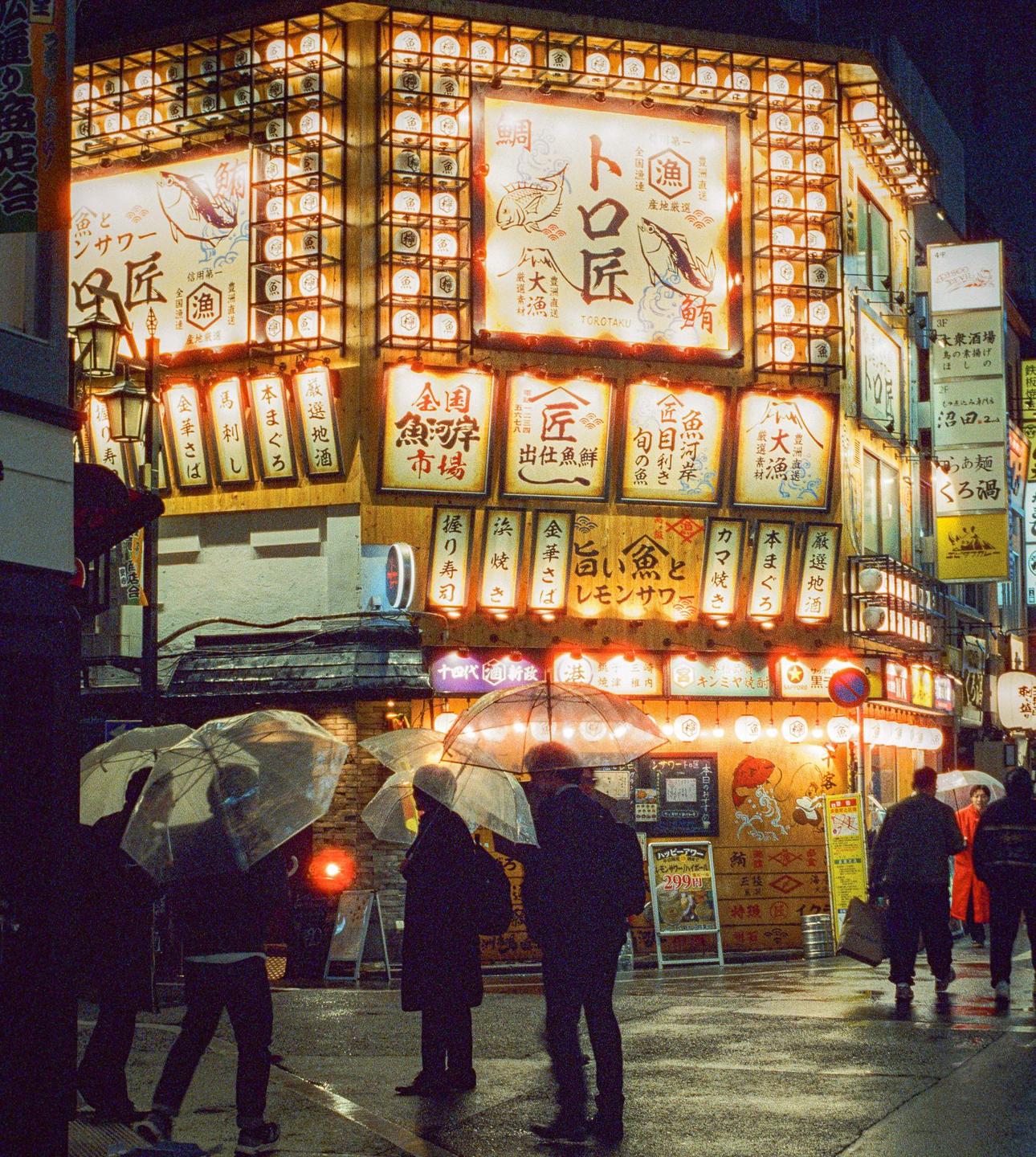 People with umbrellas walk on a wet street in front of a brightly lit building with Japanese signs at night.