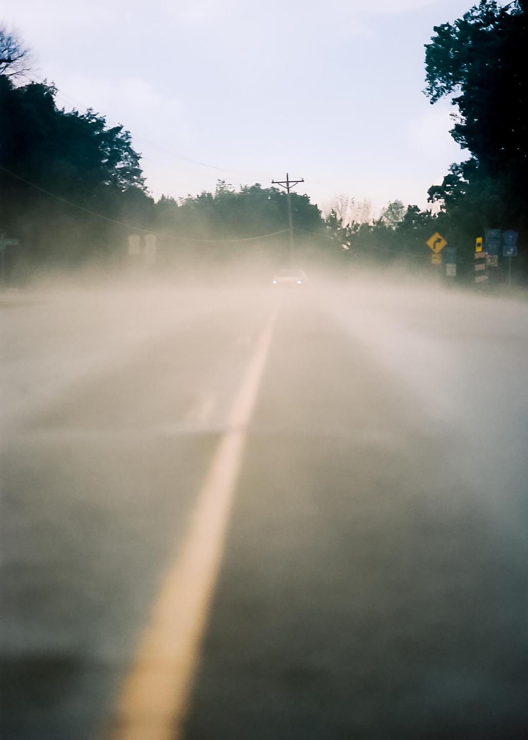 A car with its headlights on drives down a hazy road with a yellow dividing line.