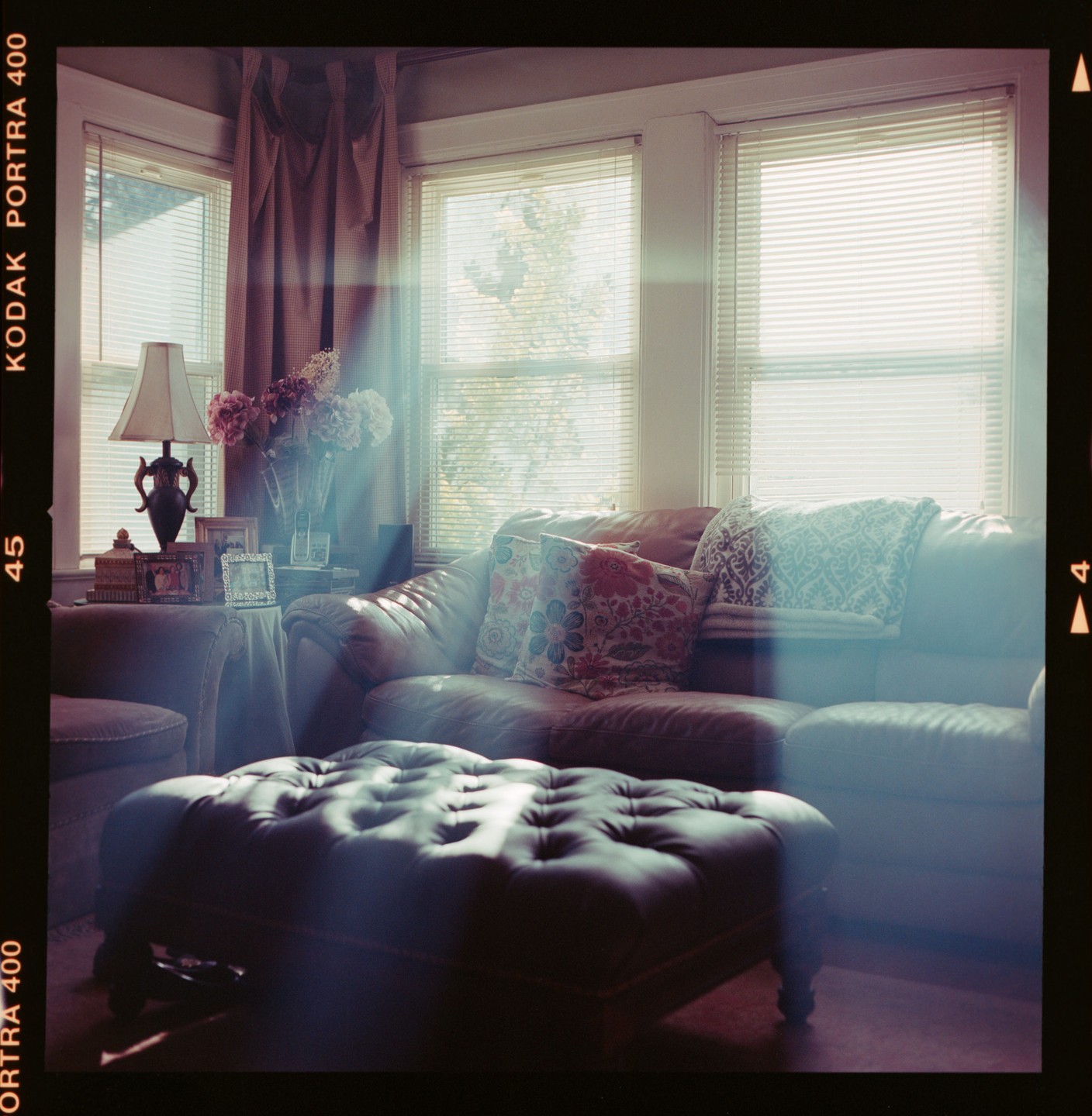 Sunlight streams through windows into a living room with a leather sofa, ottoman, and lamp.