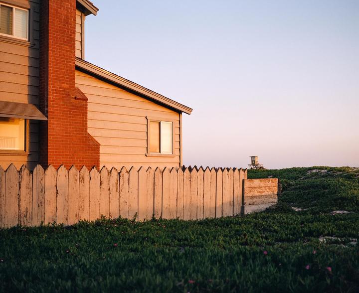 A house with a red brick chimney and a white picket fence stands next to a grassy dune with a lifeguard stand in the distance.