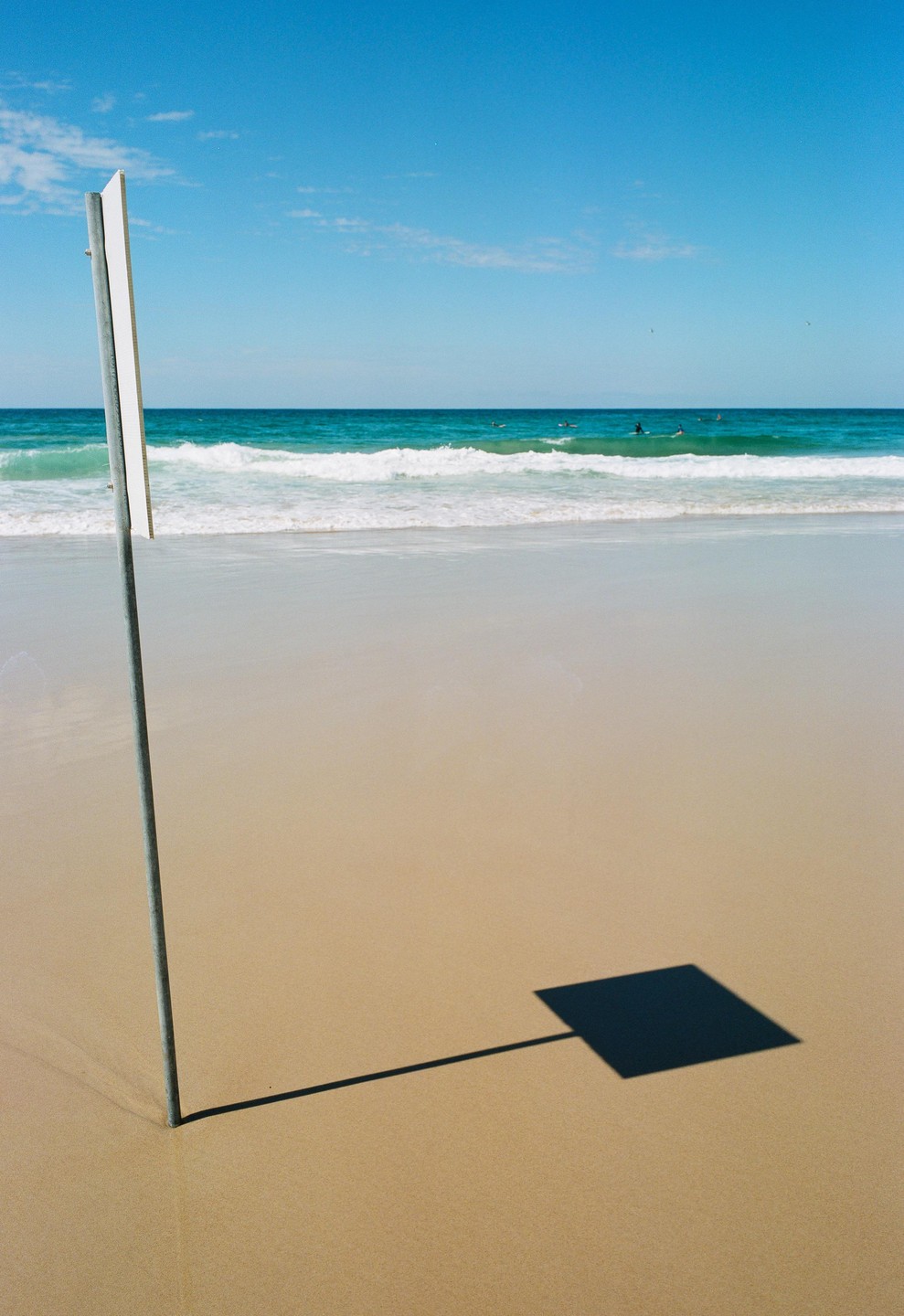 A metal pole casts a square shadow on a sandy beach with ocean waves and surfers in the background.