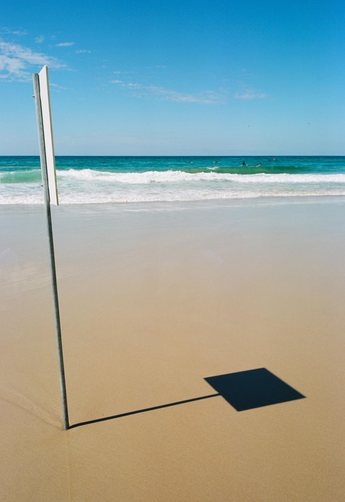 A metal pole casts a square shadow on a sandy beach with ocean waves and surfers in the background.