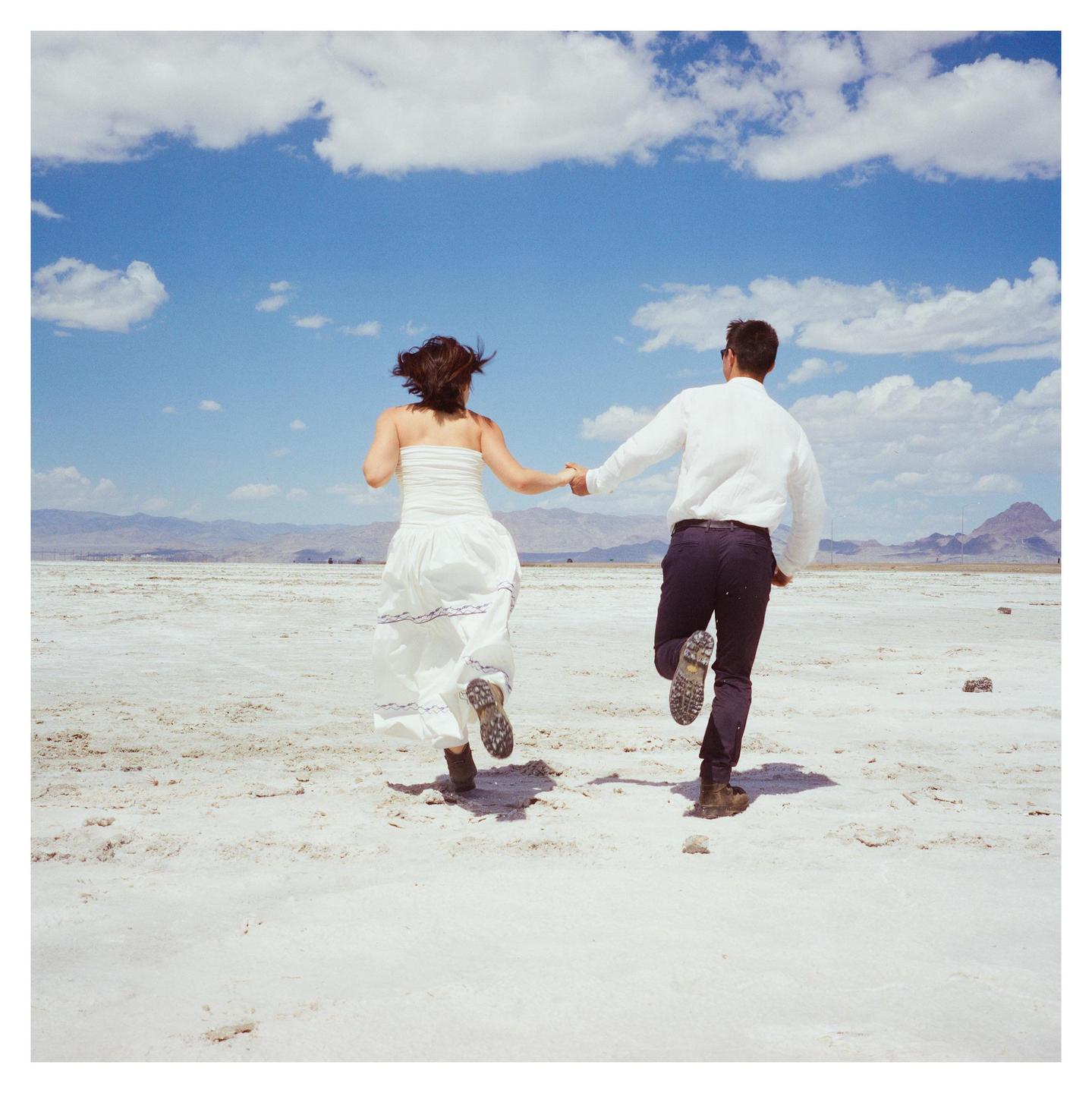 A couple runs hand-in-hand across a vast, arid landscape under a bright blue sky with scattered clouds.