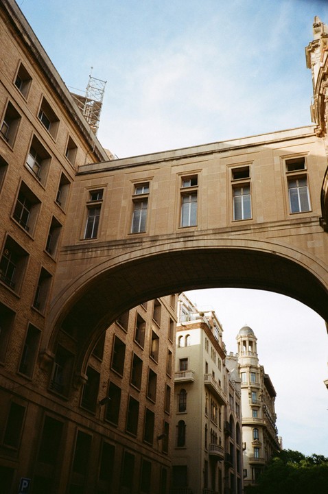 A stone archway connects two buildings under a bright sky in Barcelona.