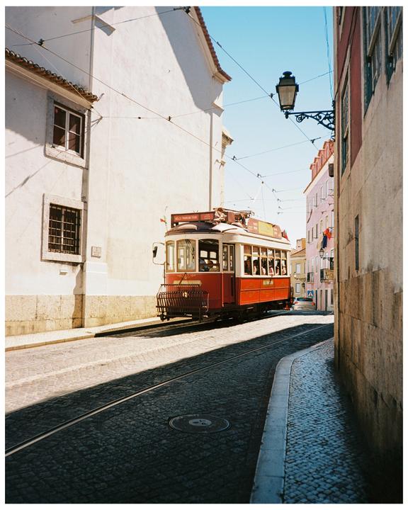 A red tram travels down a narrow cobblestone street lined with buildings.