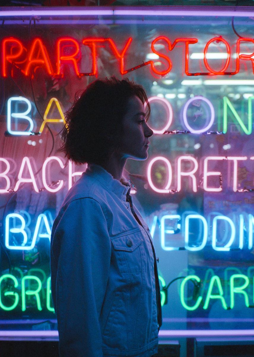 A woman in a denim jacket stands in profile against a background of colorful neon signs.