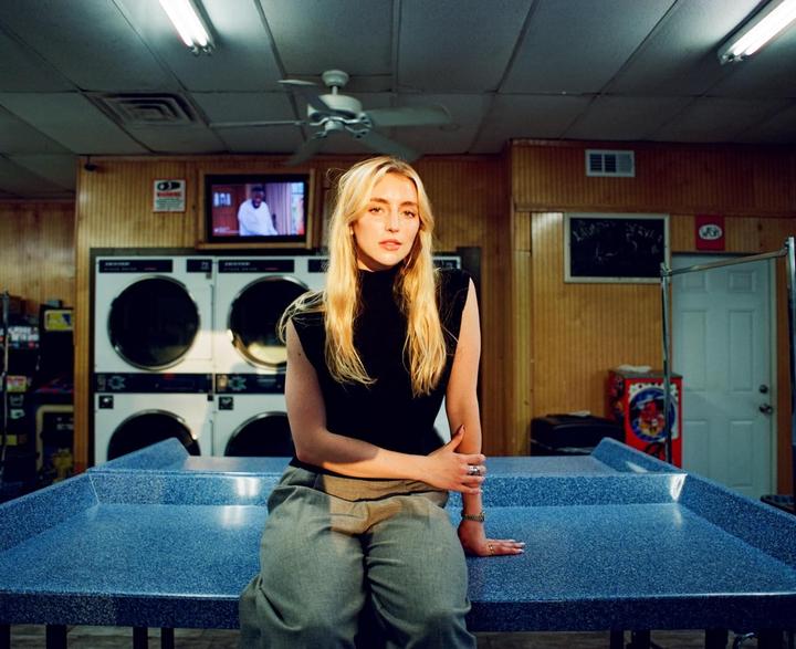 A woman sits on a blue table in a laundromat with washing machines in the background.
