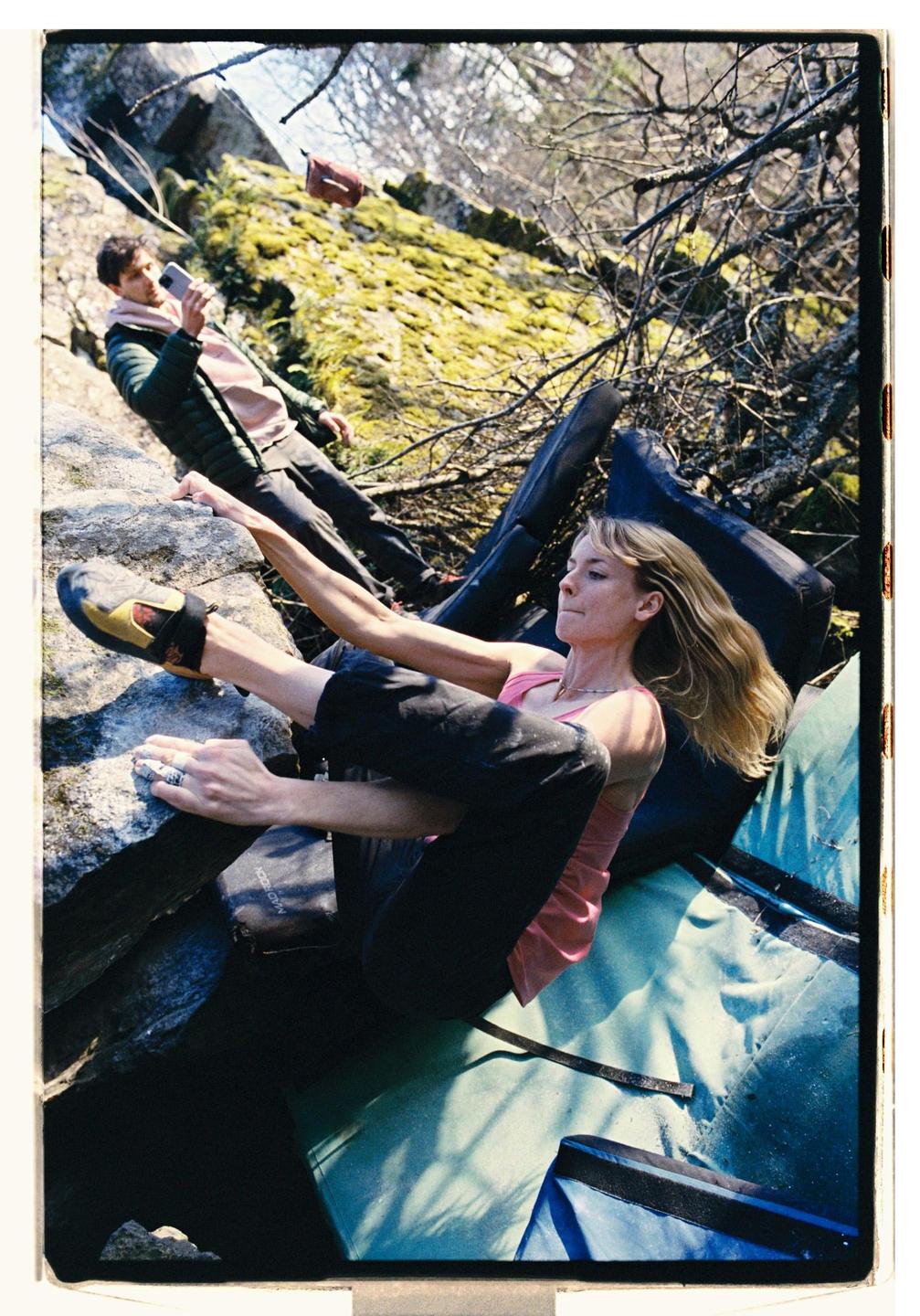 A woman bouldering on a rock with a man filming her in the background.