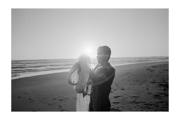 A man stands on a beach holding a fishing net with the sun setting over the ocean in the background.