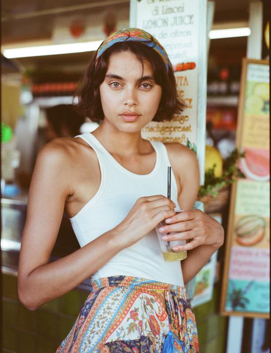 A woman with a patterned headband and skirt holds a drink with a straw in front of a juice stand.