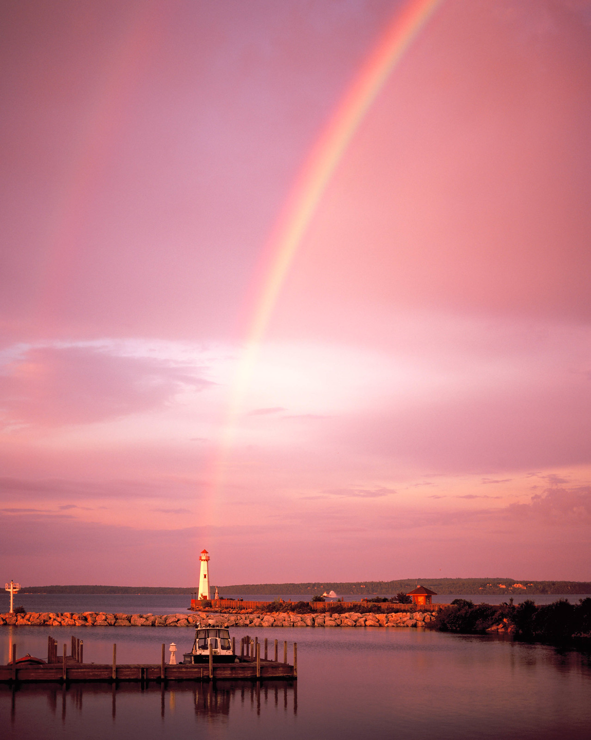 A rainbow arches over a lighthouse and a boat docked in a harbor at sunset.