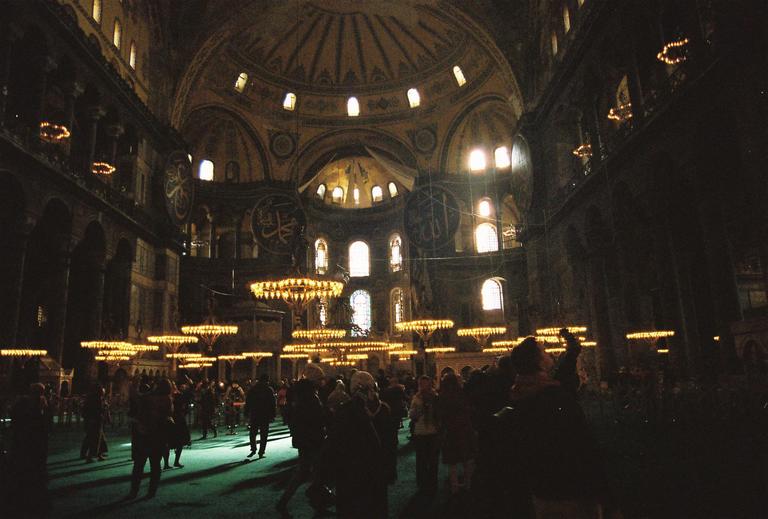 A large crowd of people stands under many chandeliers in a vast, dimly lit hall with high arched ceilings and windows.