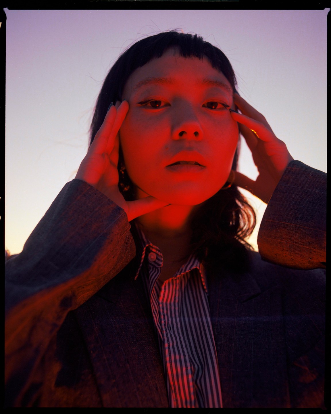 A woman with dark hair and bangs poses with her hands on her temples, illuminated by red light.