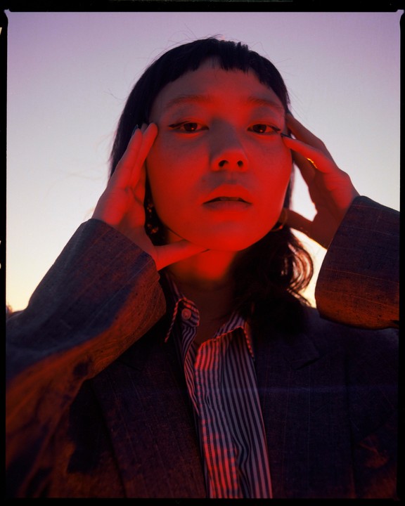 A woman with dark hair and bangs poses with her hands on her temples, illuminated by red light.