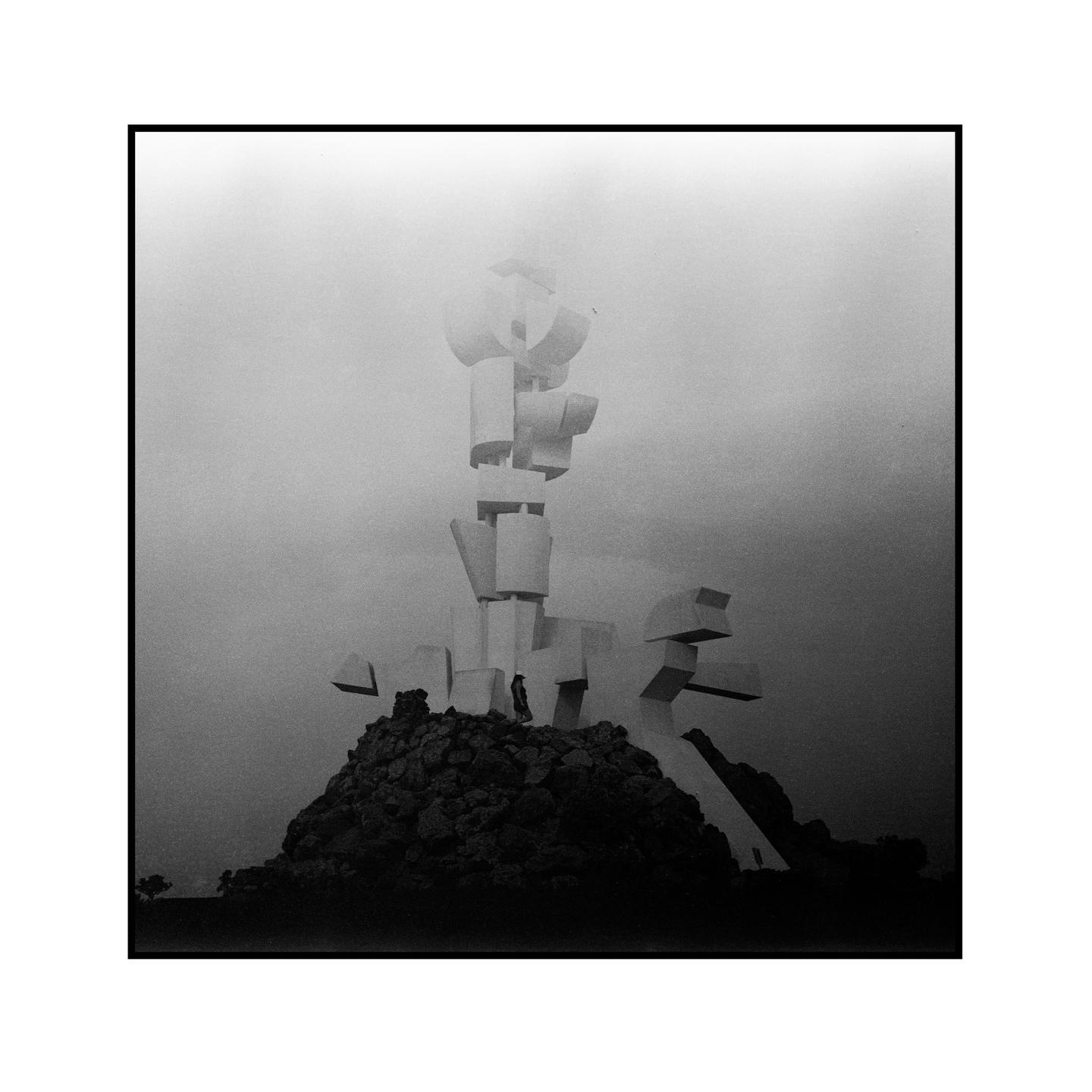 A person stands on a rocky mound in front of a large, abstract sculpture under a hazy sky.