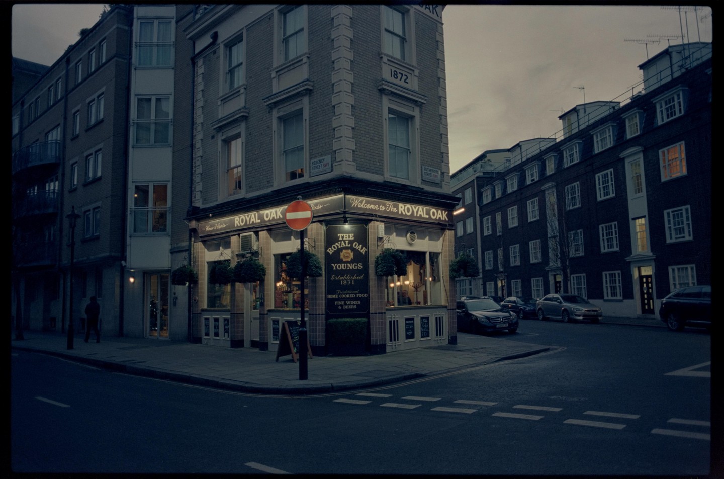 A pub with illuminated signs stands on a street corner at dusk, with cars parked along the street and a person walking in the distance.