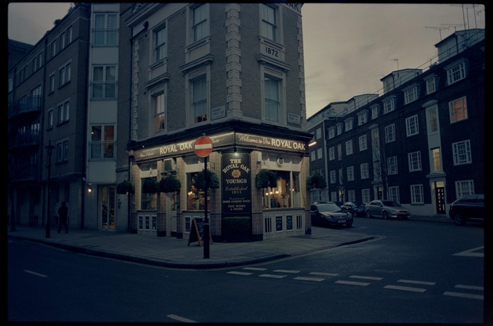 A pub with illuminated signs stands on a street corner at dusk, with cars parked along the street and a person walking in the distance.